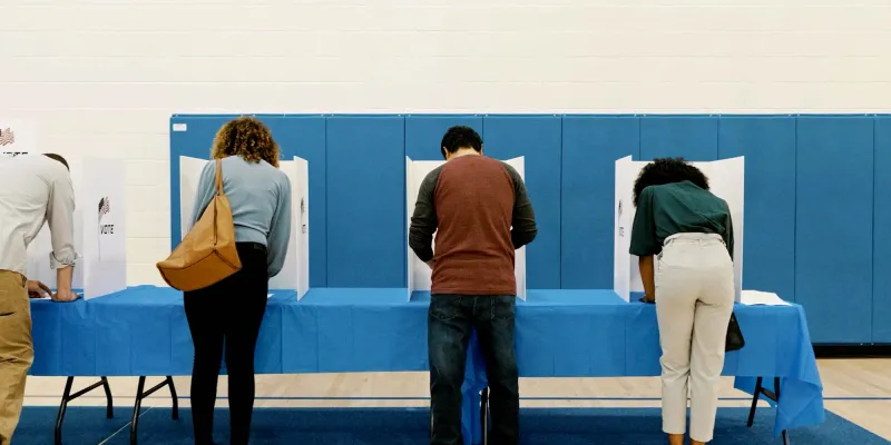 People casting votes at a polling station with blue partitions and a row of empty chairs.