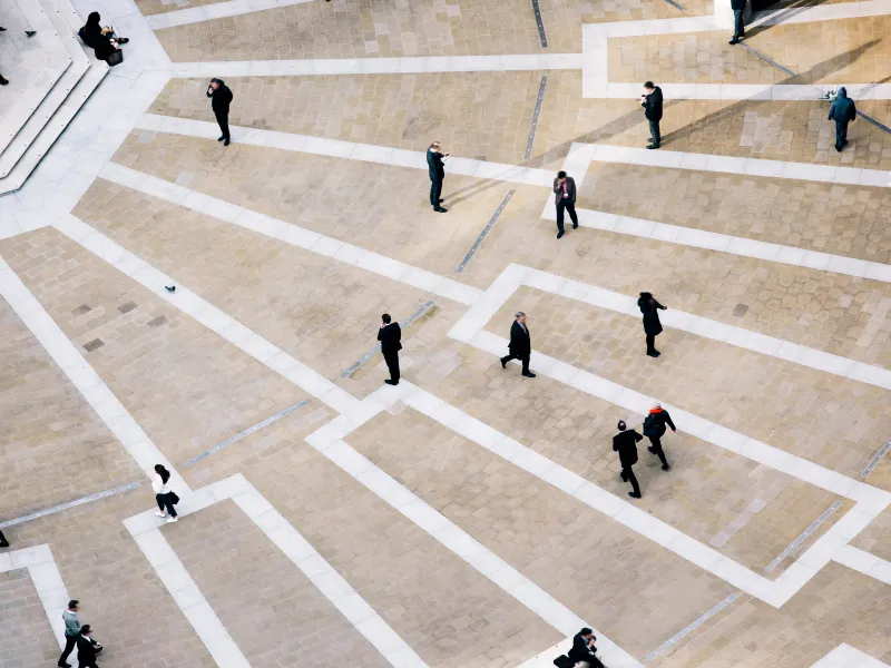 People walking across a large, patterned paved plaza with white lines and tiles.