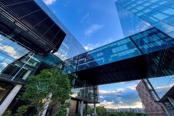 Modern glass buildings with skybridge and trees beneath, reflecting blue sky.